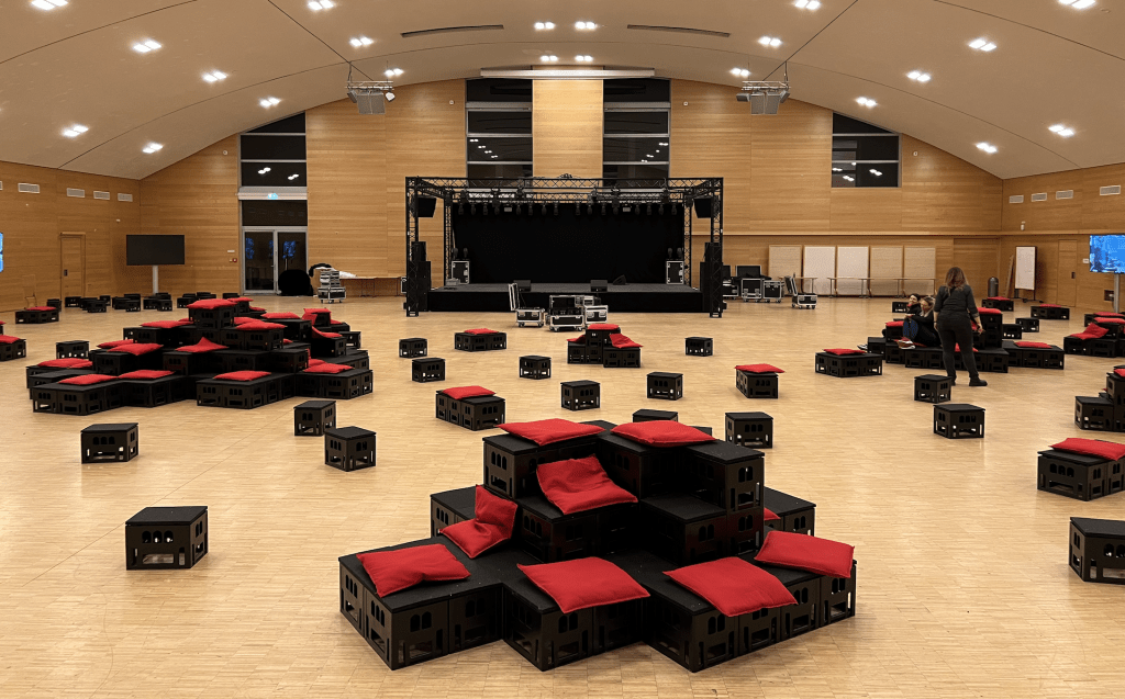 Veranstaltungssaal mit pyramidenförmig arrangierten cratedesign®-Kisten, die mit roten Kissen ausgestattet sind.